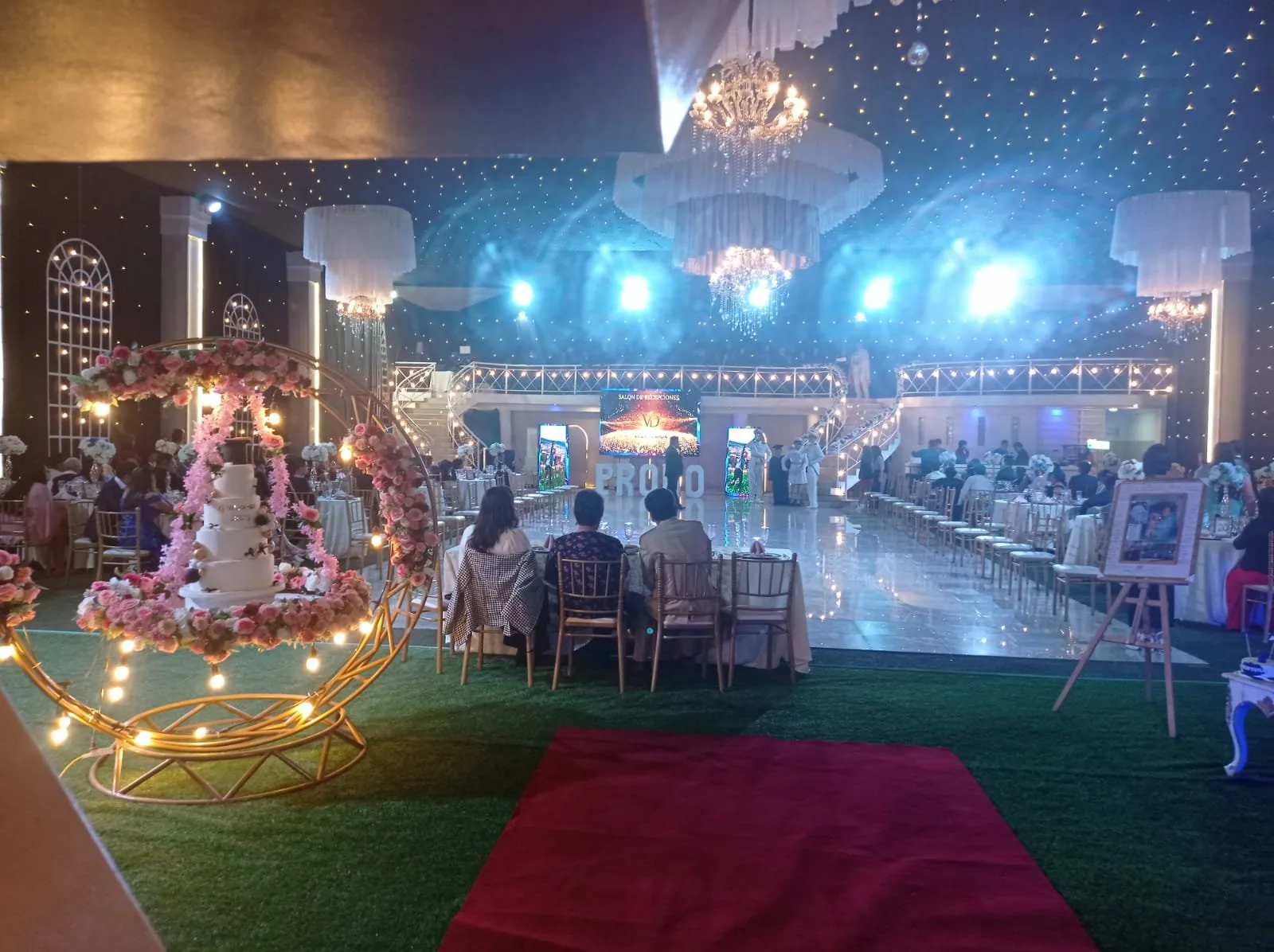 Pastel de celebración sobre soporte floral, con luces en forma de corazón al fondo